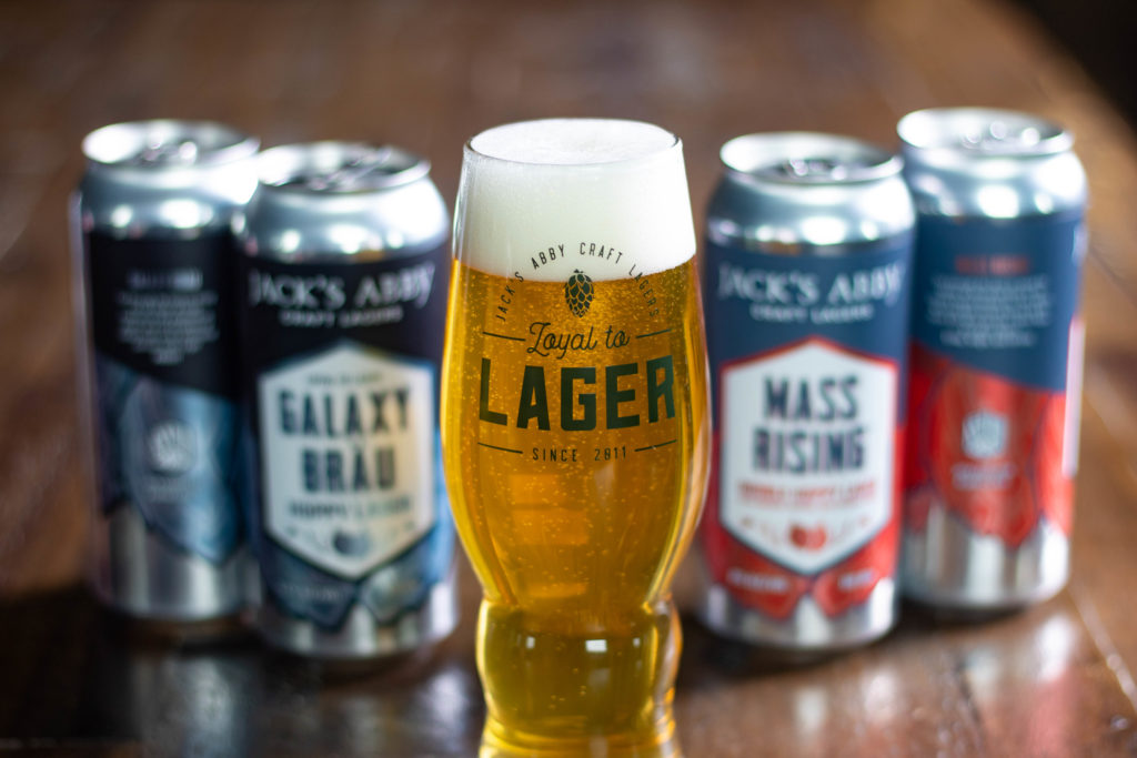 Close up photo of a clear beer in glass, with cans of Jack's Abby in background.